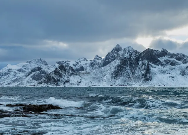 Bølger slår mot kysten foran snødekte fjell under tung himmel og dramatisk lys.